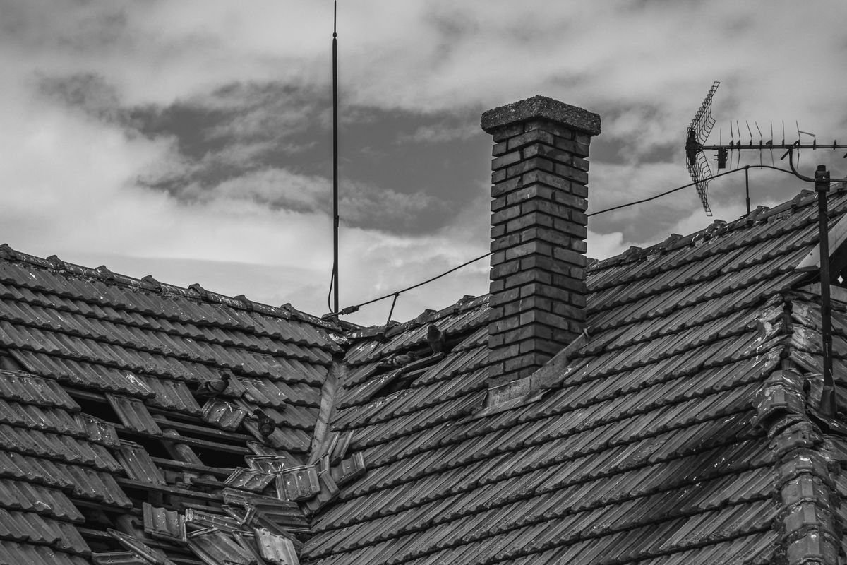 Damaged roof with broken tiles around a chimney under an overcast sky
