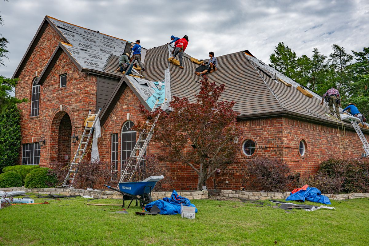Roofing crew handling shingles and materials on the roof of a brick house